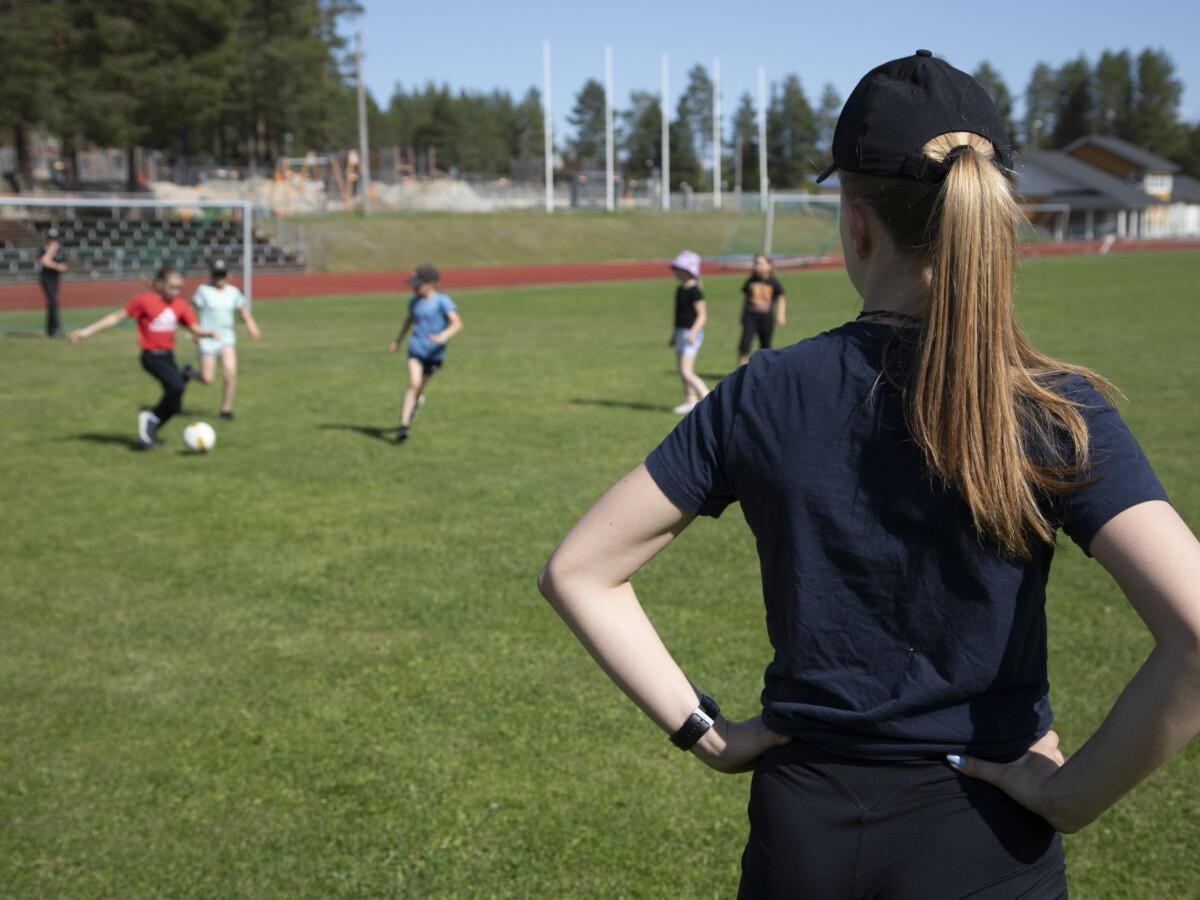 nainen seisoo jalkapallokentällä, taustalla lapset pelaavat jalkapalloa