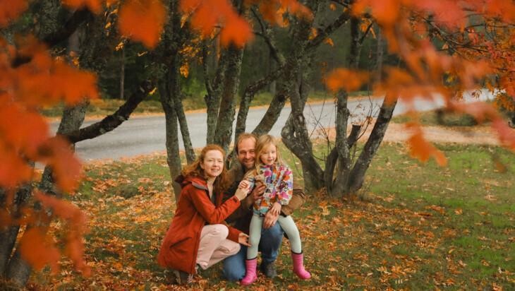 Isä, äiti ja lapsi hymyilevät, syksyinen maisema